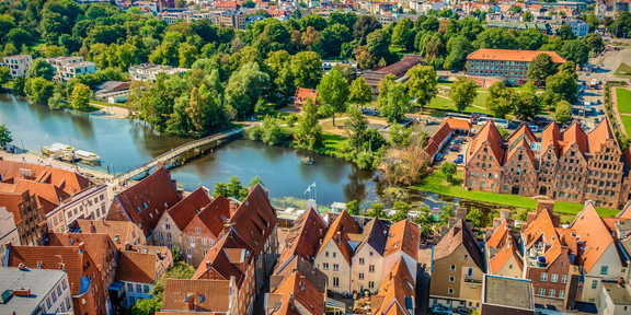 Lübeck (Modul 7) Lübeck aus der Vogelperspektive auf der man Wasser, Haussiedlungen und Wälder sehen kann.