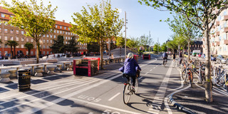 Es ist eine Straße mit zwei Fahrradfahrern umgeben von Häusern, Bäumen, stehenden Fahrrädern und Sitzgelegenheiten.