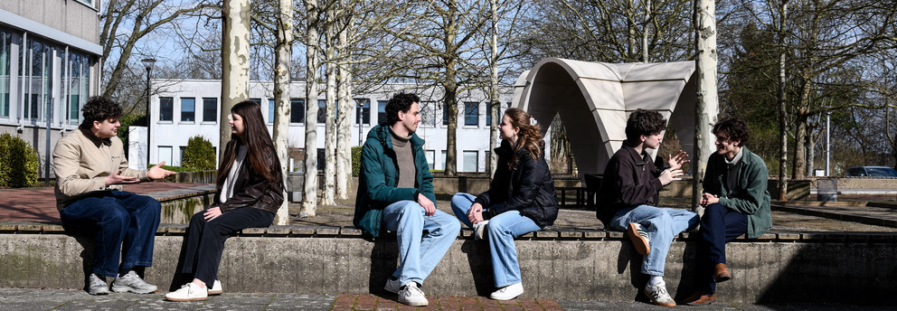 Studierende sitzen auf einer Sitzfläche am Campus und unterhalten sich.