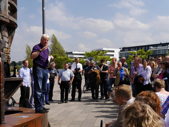 Menschengruppe versammelt sich im Halbkreis vor dem Redner an einem Denkmal.