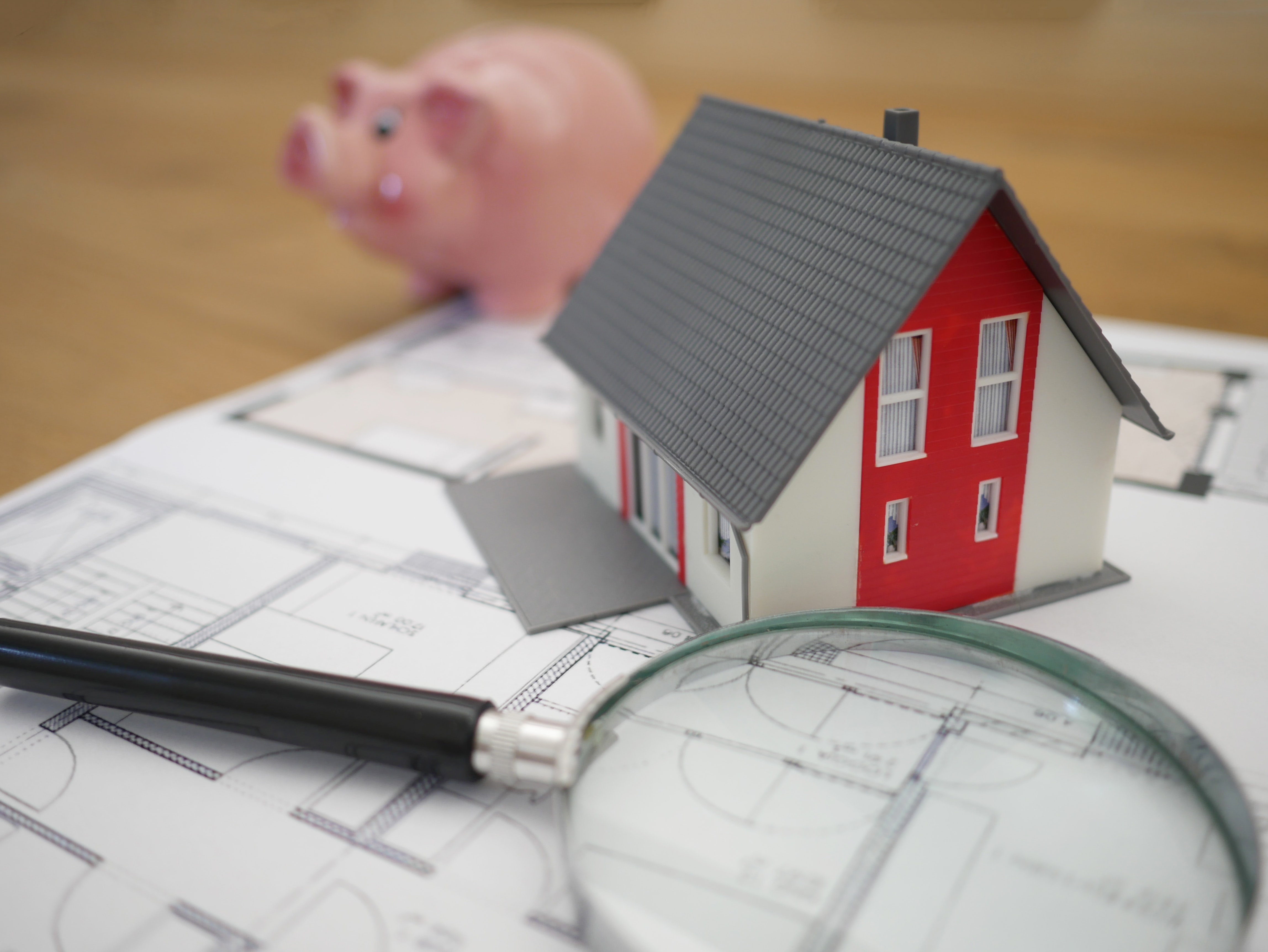 Ein Modellhaus steht auf einem Bauplan mit einer Lupe und einem Sparschwein daneben.