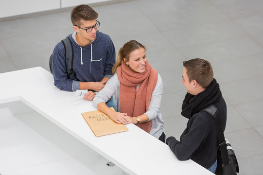 Three people are standing in front of a service desk. The person in the middle is holding an envelope.
