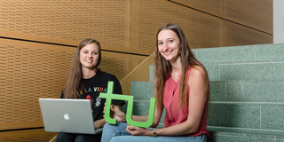 Students with laptop and TU logo_02 Two students are sitting on a stair. One is holding a laptop and the other the logo of TU Dortmund University