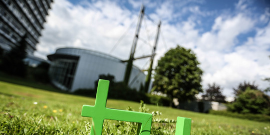 Im Vordergrund steht das TU-Logo im Gras, im Hintergrund das Audimax.