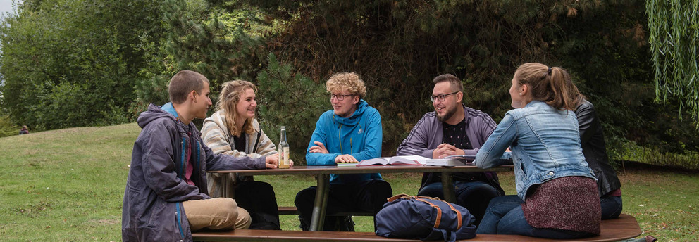 Sechs Studierende sitzen an einer Sitzgruppe auf der Wiese mit Bäumen im Hintergrund. 