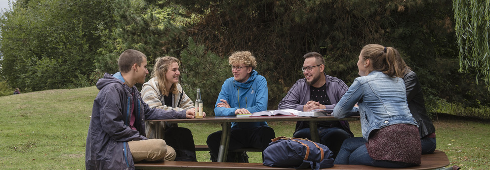 Sechs Studierende sitzen an einer Sitzgruppe auf der Wiese mit Bäumen im Hintergrund. 