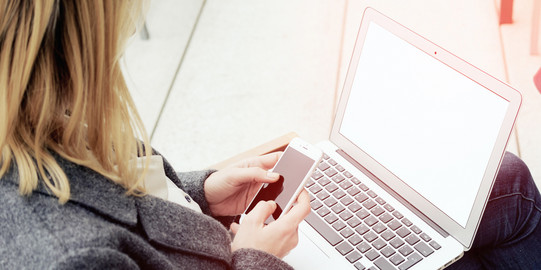 Woman with laptop on her lap, holding a smartphone in her hands.