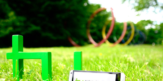 TU-App Ein Smartphone, das neben dem TU-Logo im Gras steht, im Hintergrund sind die Spektralringe zu sehen.