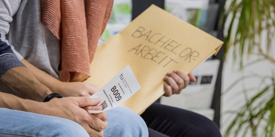 Close-up of two people sitting nect to each other. One is holding an envelope, the other a small piece of paper with a number printed on it