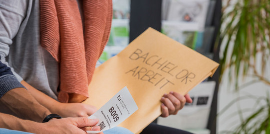 Close-up of two people sitting nect to each other. One is holding an envelope, the other a small piece of paper with a number printed on it