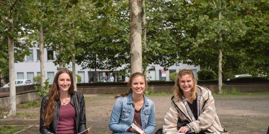 Drei Studentinnen sitzen auf einer Bank, halten Bücher in ihren Händen und lächeln.
