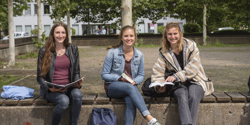 Drei Studentinnen sitzen auf einer Bank, halten Bücher in ihren Händen und lächeln.