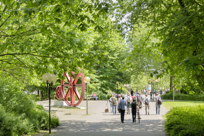 Students walk past the cogwheel and green trees line the path.