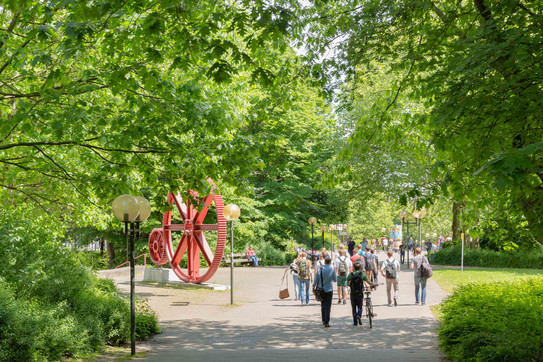 TU Dortmund im Sommer_04 Studierende laufen am Zahnrad vorbei und grüne Bäume säumen den Weg.