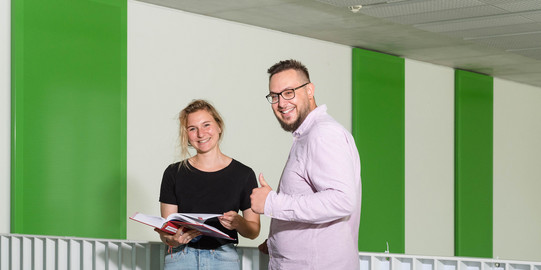 Zwei Studierende stehen vor einem Geländer, einer streckt den Daumen hoch.