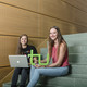 Two students are sitting on a stair. One is holding a laptop and the other the logo of TU Dortmund University