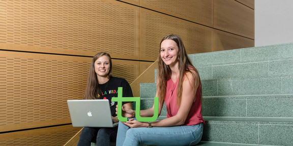 Students with laptop and TU logo_03 Two students are sitting on a stair. One is holding a laptop and the other the logo of TU Dortmund University
