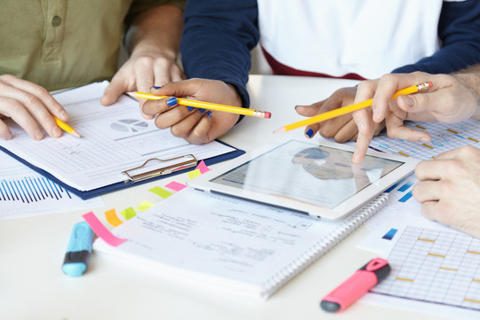 On a table are leaves and blocks, a tablet that is used with one hand. You can see hands with pencils and markers.  Charts and tables can be seen on the papers.