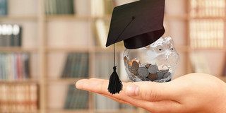 A transparent piggy bank full of coins and with a doctoral cap is held in front of a book shelf. 